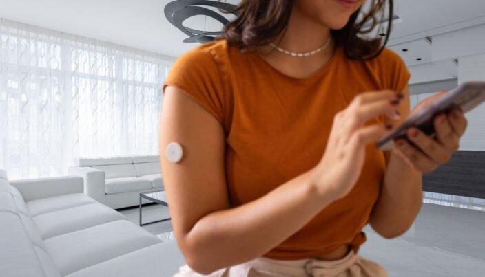 Medidor de Glicose no Braço Oferecendo Conforto e precisão 1 Pessoa sentada utilizando um medidor de glicose no braço enquanto monitora os níveis de açúcar no sangue em um smartphone.